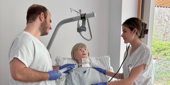 Zwei Personen stehen an einem Krankenbett und üben an einer Übungspuppe medizinische Tätigkeiten