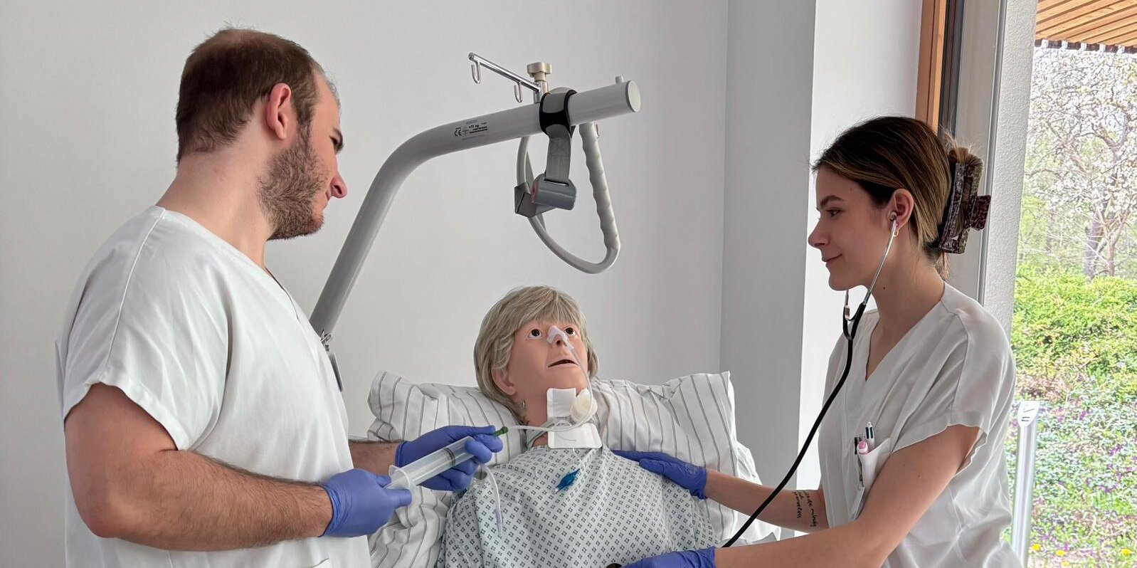 Zwei Personen stehen an einem Krankenbett und üben an einer Übungspuppe medizinische Tätigkeiten