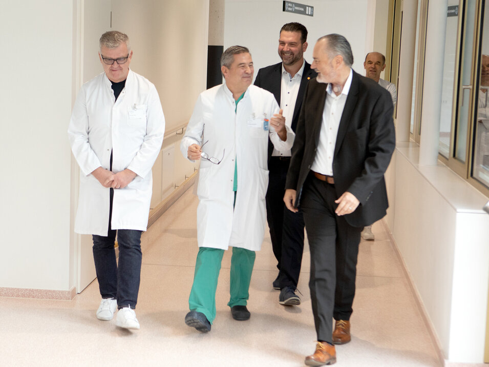 Drei Personen auf dem Weg zu einer Pressekonferenz, im Hintergrund eine vierte Person