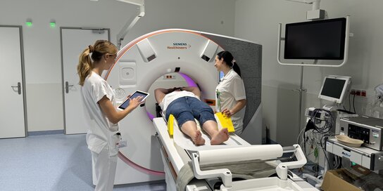 Zwei weibliche Personen in Krankenhauskleidung stehen vor einem MR-Gerät, in dem eine Patientin liegt