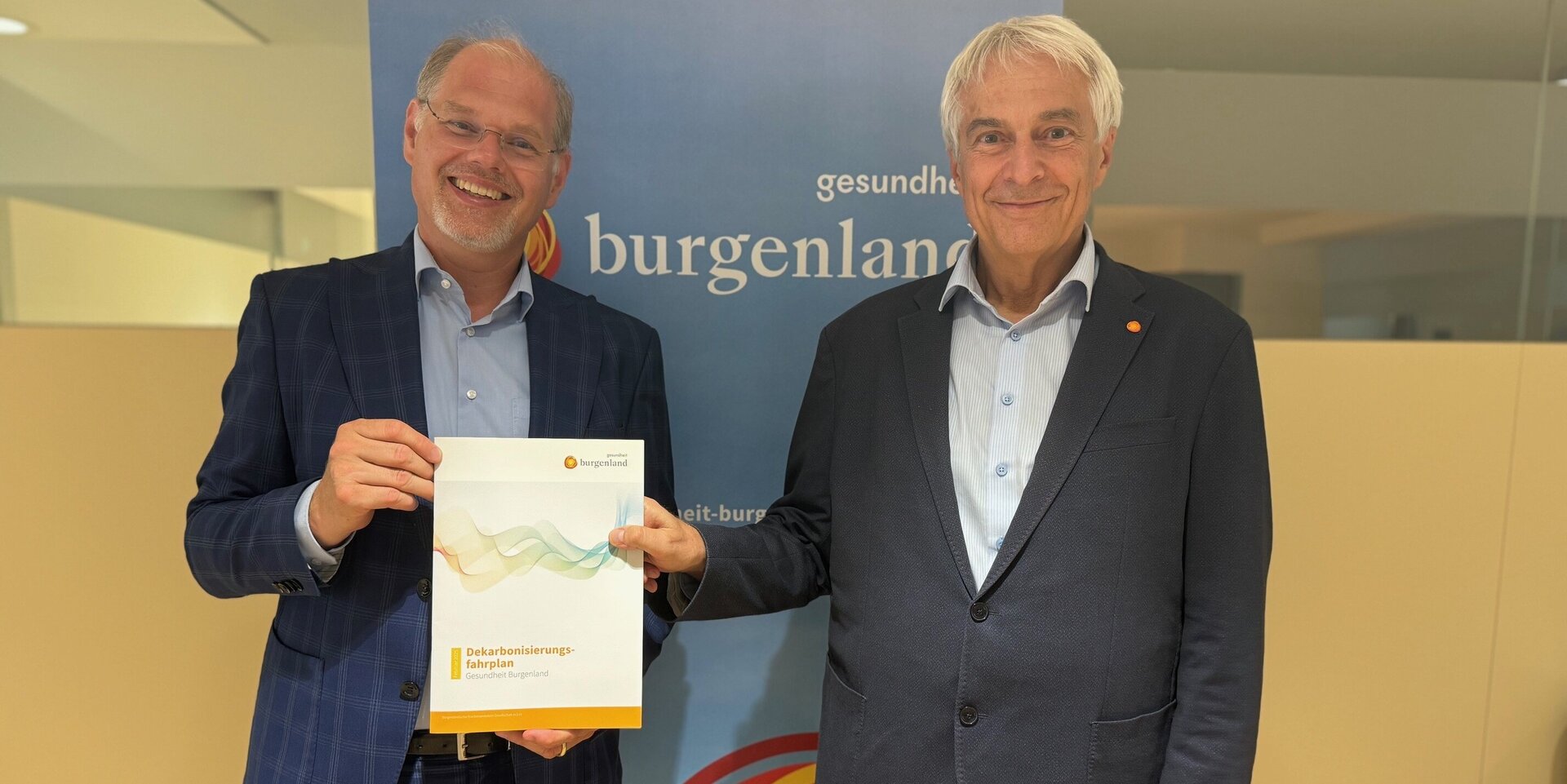 Foto: Gesundheit Burgenland Zwei männliche Personen im Anzug stehen nebeneinander, einer hält einen Umweltbericht in der Hand