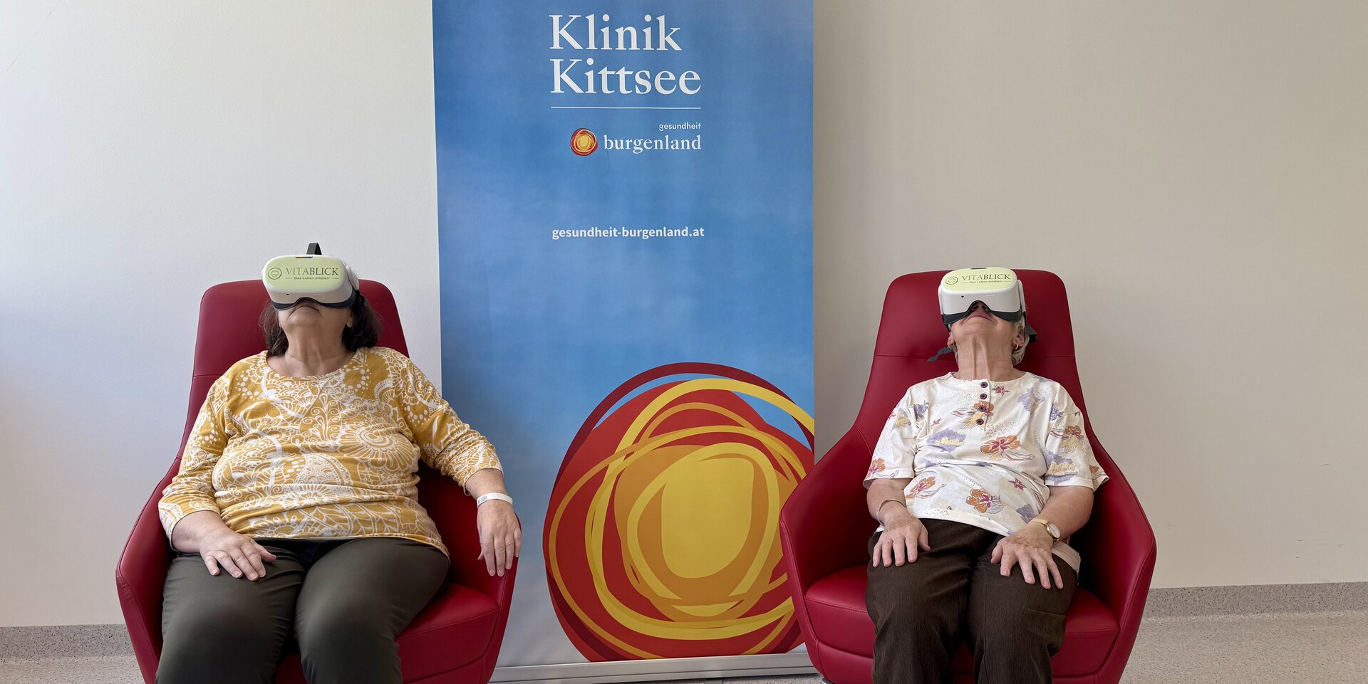 Foto: Gesundheit Burgenland Zwei Frauen sitzen vor einem Roll-up mit VR-Brillen in zwei roten Stühlen