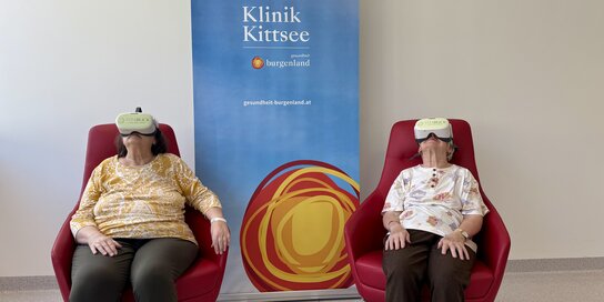 Zwei Frauen sitzen vor einem Roll-up mit VR-Brillen in zwei roten Stühlen