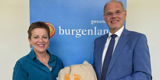 Eine Frau und ein Mann stehen vor einem Roll-Up der Gesundheit Burgenland und präsentieren ein Sackerl mit aufgedruckten Babyfüßen