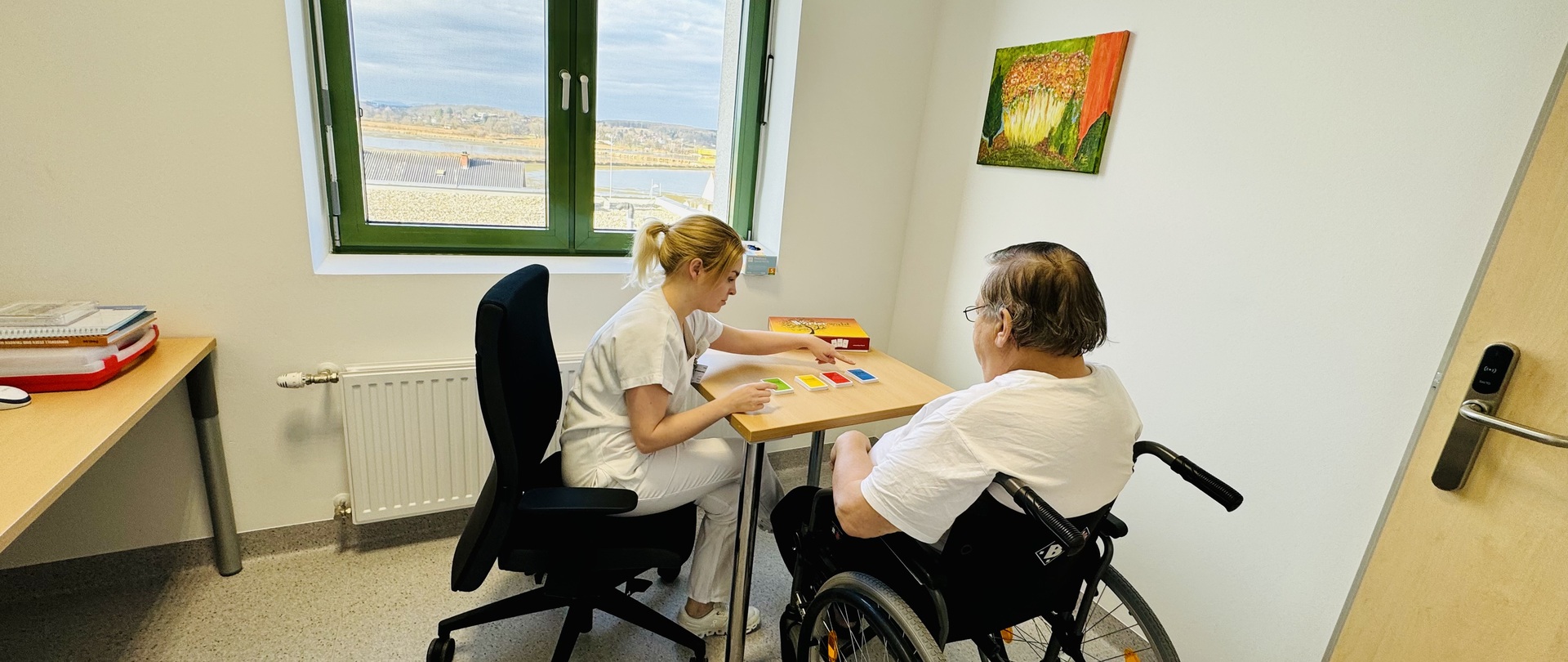 Logopädin Verena Schaffler während einer Therapiesitzung mit einem Patienten im Rollstuhl, auf dem Tisch liegen Karten in verschiedenen Farben