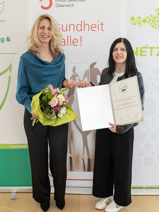 Zwei Frauen, eine mit einer Auszeichnung in Papierform, eine mit Blumenstrauß