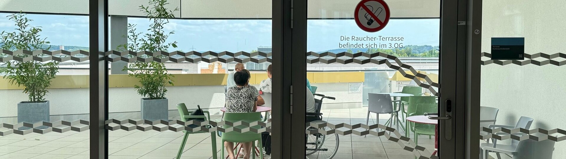 Foto: Gesundheit Burgenland  Blick auf eine Terrasse, auf der Tische und Sessel stehen und drei Menschen sitzen