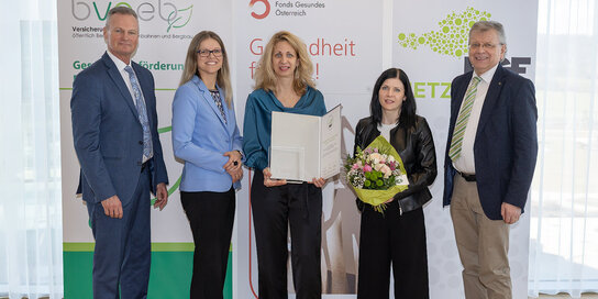 Fünf Personen stehen in einer Linie, eine hält einen Blumenstrrauß, eine andere eine Auszeichnung in Papierform