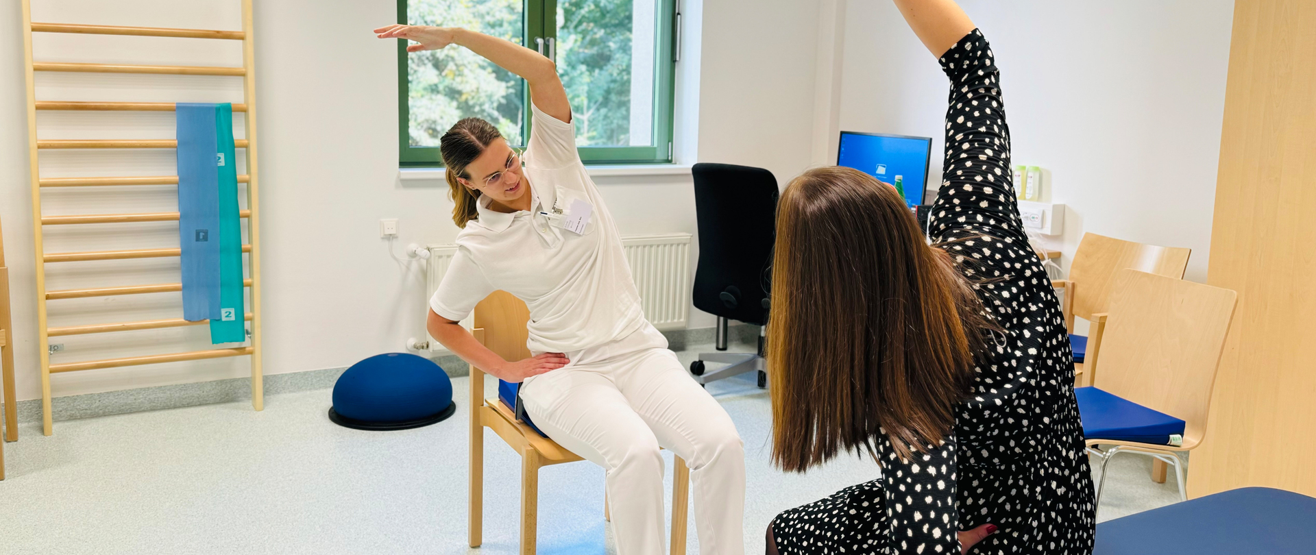 Eine Physiotherapeutin zeigt einer Patientin eine Übung vor, die diese nachmacht