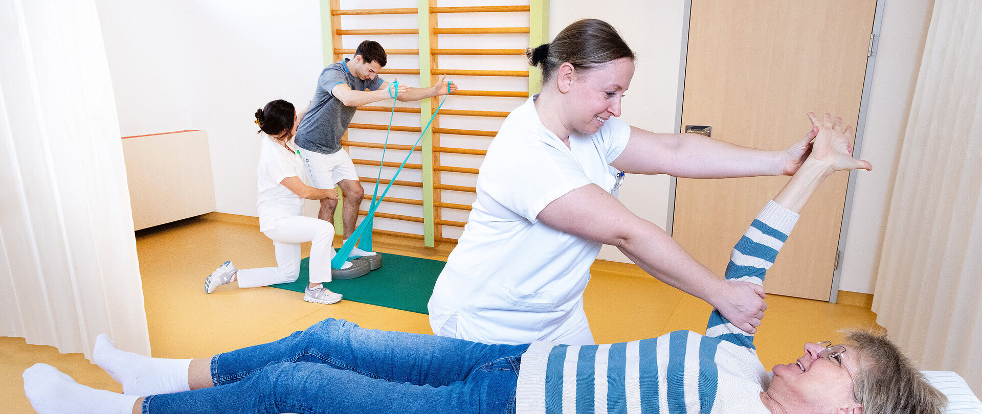 Foto: Carmen Neumann-Strobl Therapieraum mit Sprossenwand: Eine Physiotherateutin macht Übung mit jungem Patienten im Stehen auf Trainingsgerät und mit Terraband. Andere Therapeutin macht Übung beim rechtem Arm einer älteren Frau, die im Bett liegt