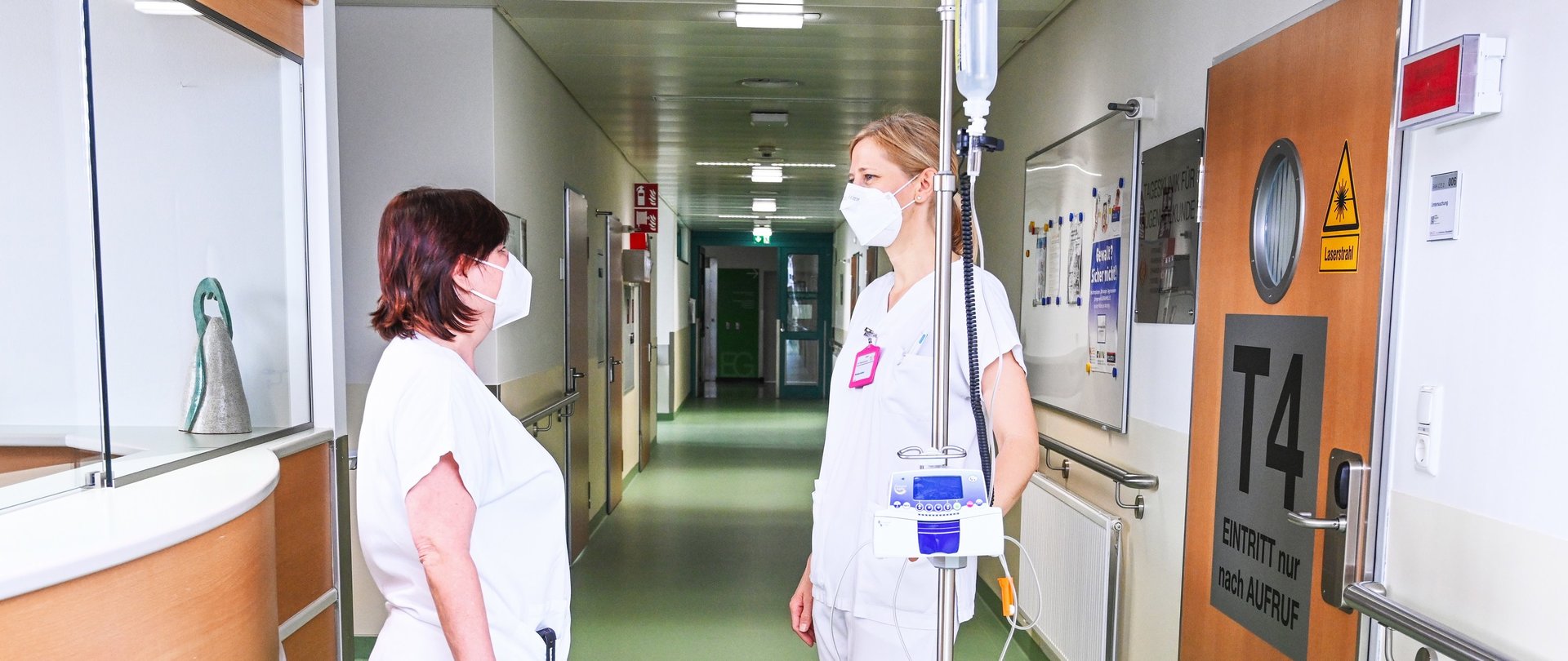 Zwei Frauen in Weiß stehen in einem Gang der Klinik, eine schiebt einen Tropf am Ständer