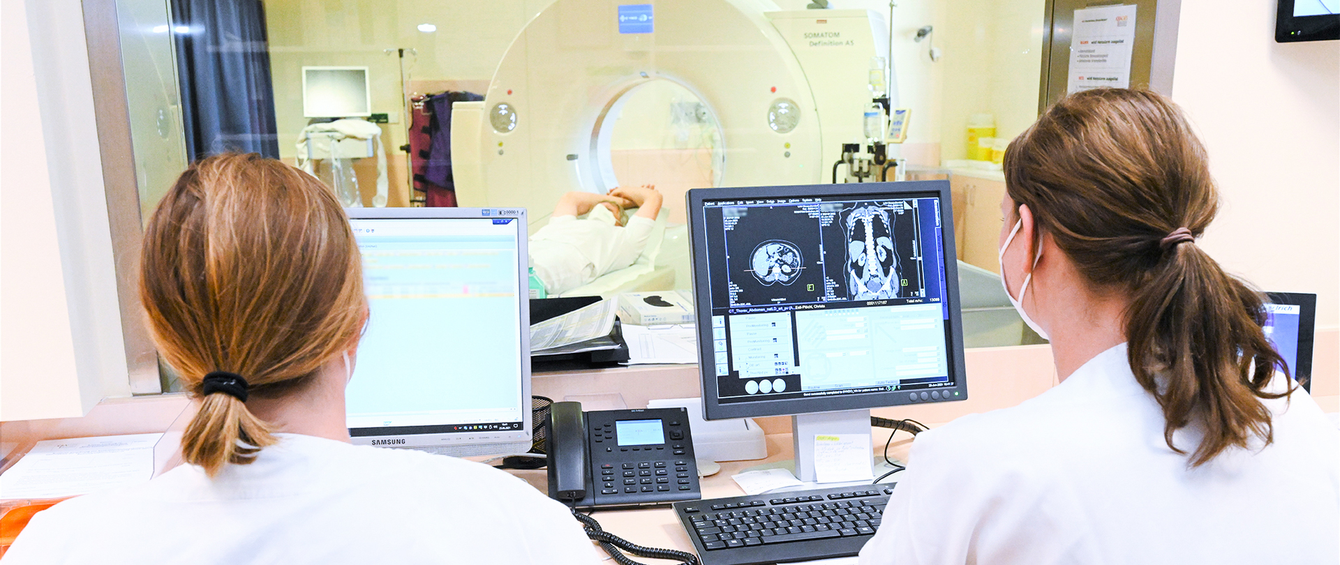 Zwei Frauen sitzen mit dem Gesicht abgewandt und blicken auf MR-Aufnahmen auf einem Bildschirm, im Hintergrund ist ein MR-Gerät mit einem Patienten zu sehen