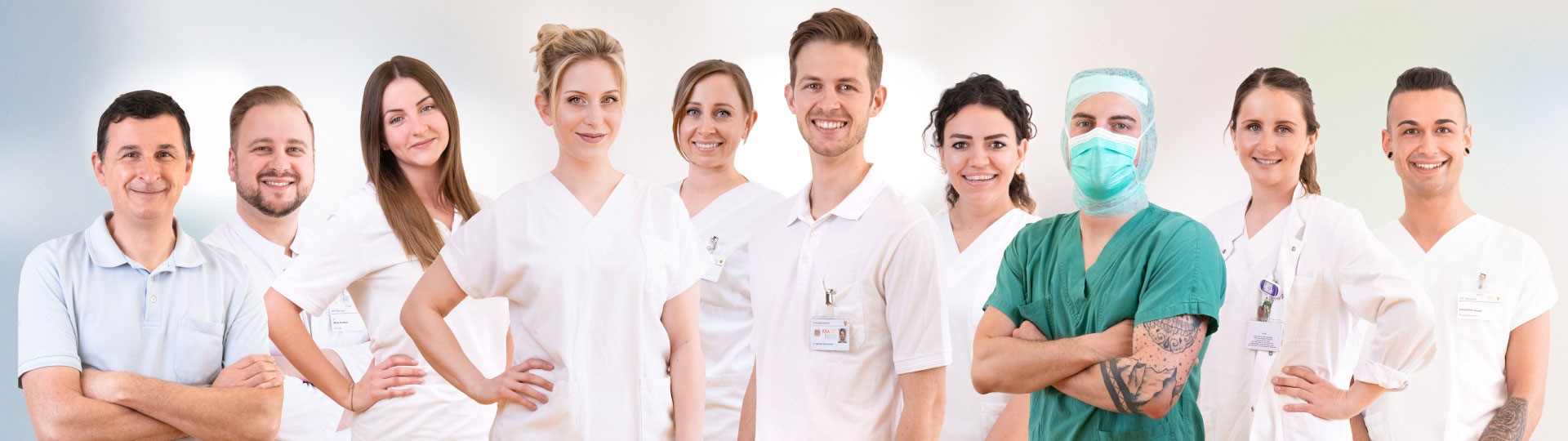 Teamfoto von verschiedenen Berufsrichtungen einer Klinik (4 weibliche und 4 männliche Personen in weißer Berufskleidung, eine männliche Person in grüner OP-Bekleidung mit Maske)